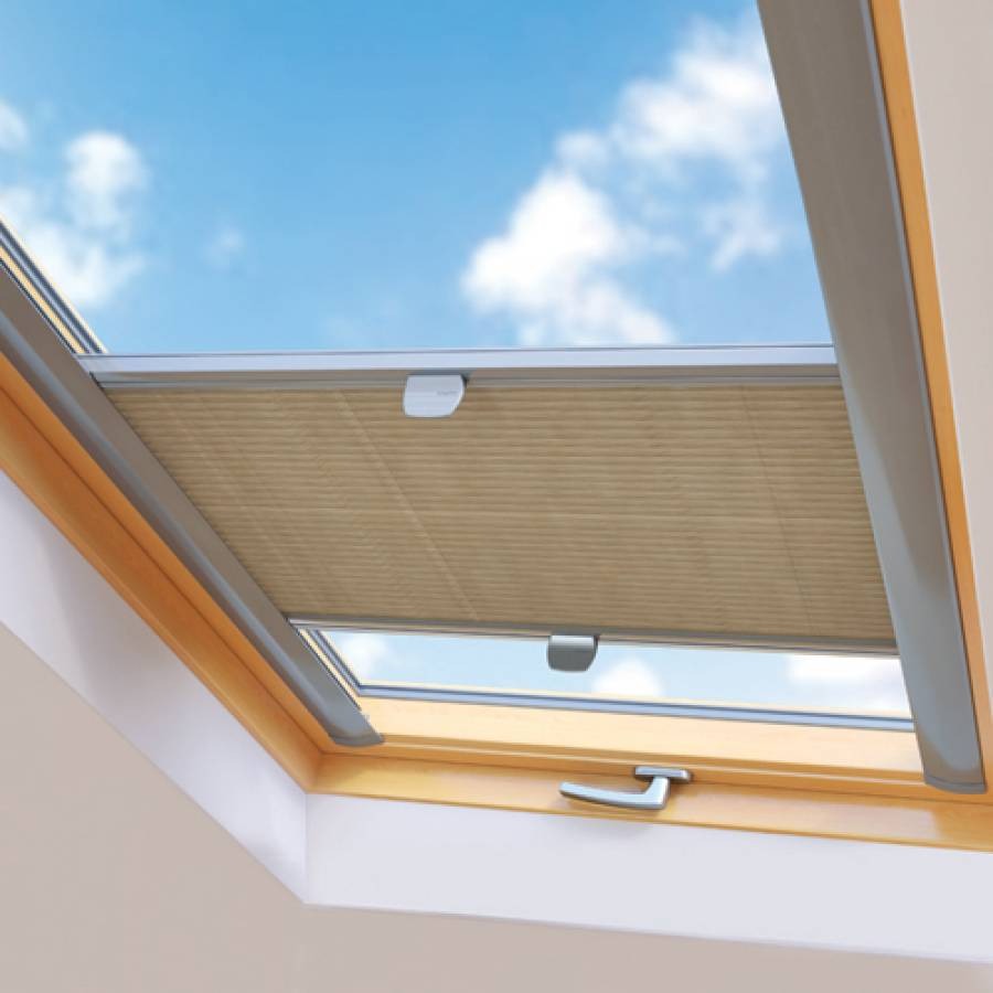 Beżowa osłona okienna plisowana, zamontowana w oknie dachowym, z widocznym uchwytem i fragmentem błękitnego nieba w tle.