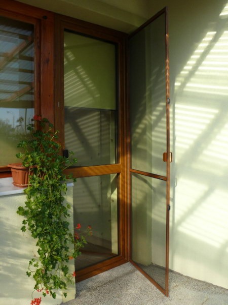 Drewniane okno z zieloną roletą wewnętrzną i otwartymi drzwiami z moskitierą; na parapecie doniczka z kwiatami.