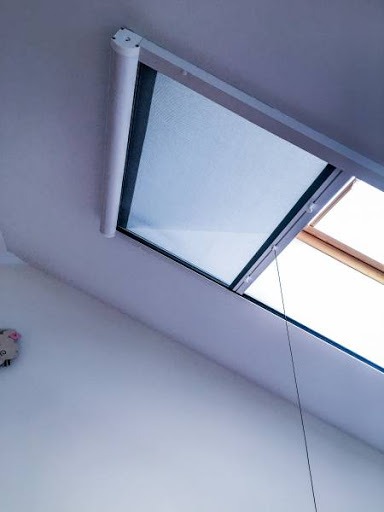 Zamknięta, jasna plisa okienna zamontowana na oknie dachowym, widoczna z dołu pod kątem, z białym mechanizmem zwijającym i cienką linką sterującą.