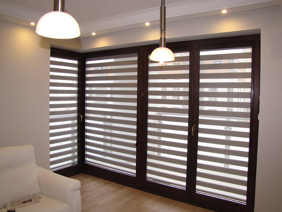 Eleganckie rolety dzień i noc w brązowych ramach okiennych, widoczne z wnętrza pokoju z białym fotelem i drewnianą podłogą, oświetlone lampami sufitowymi.