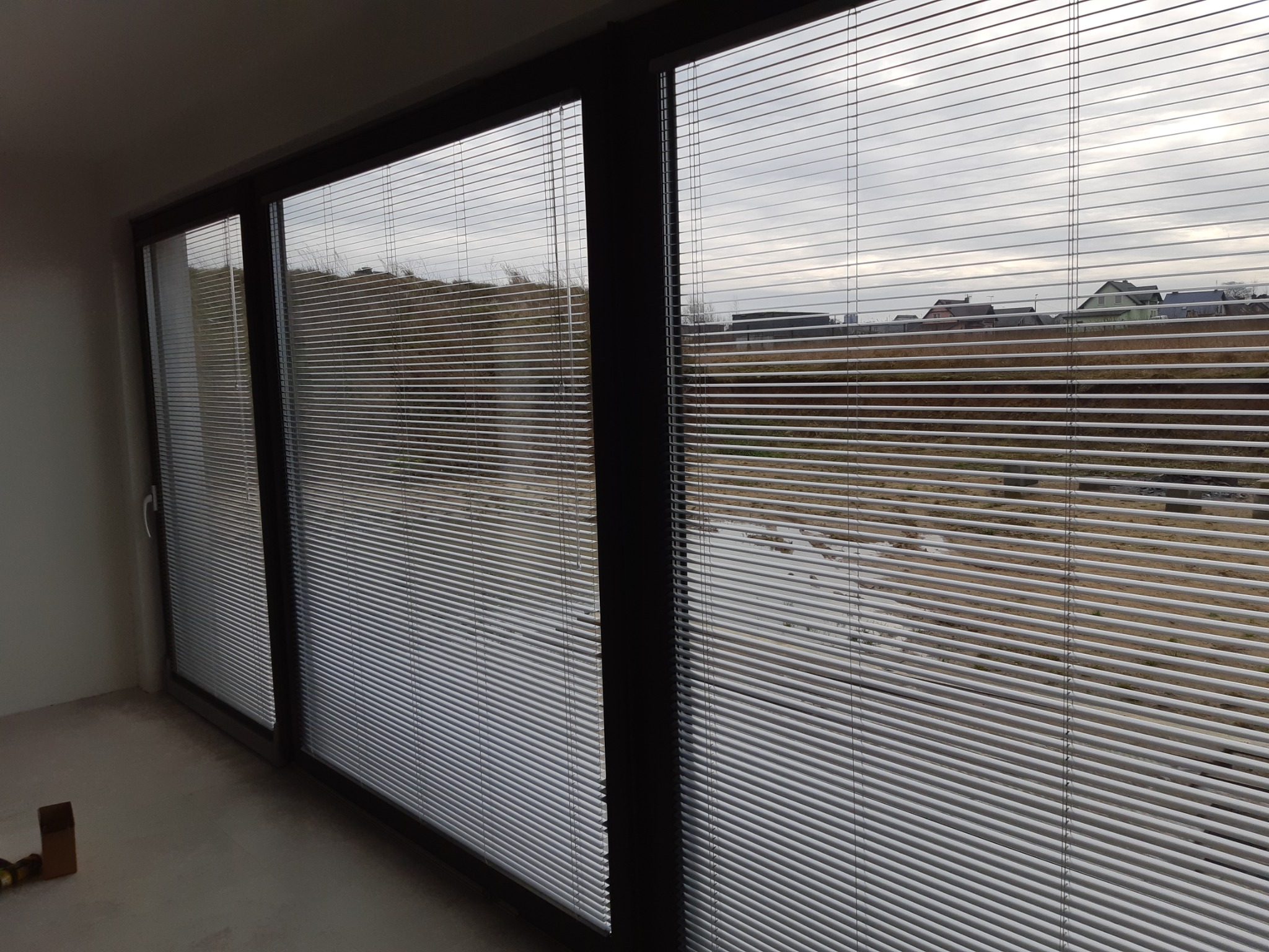 Zamknięte, poziome, aluminiowe żaluzje w oknach przesuwnych, widok na zewnątrz z szarą, pochmurną pogodą.