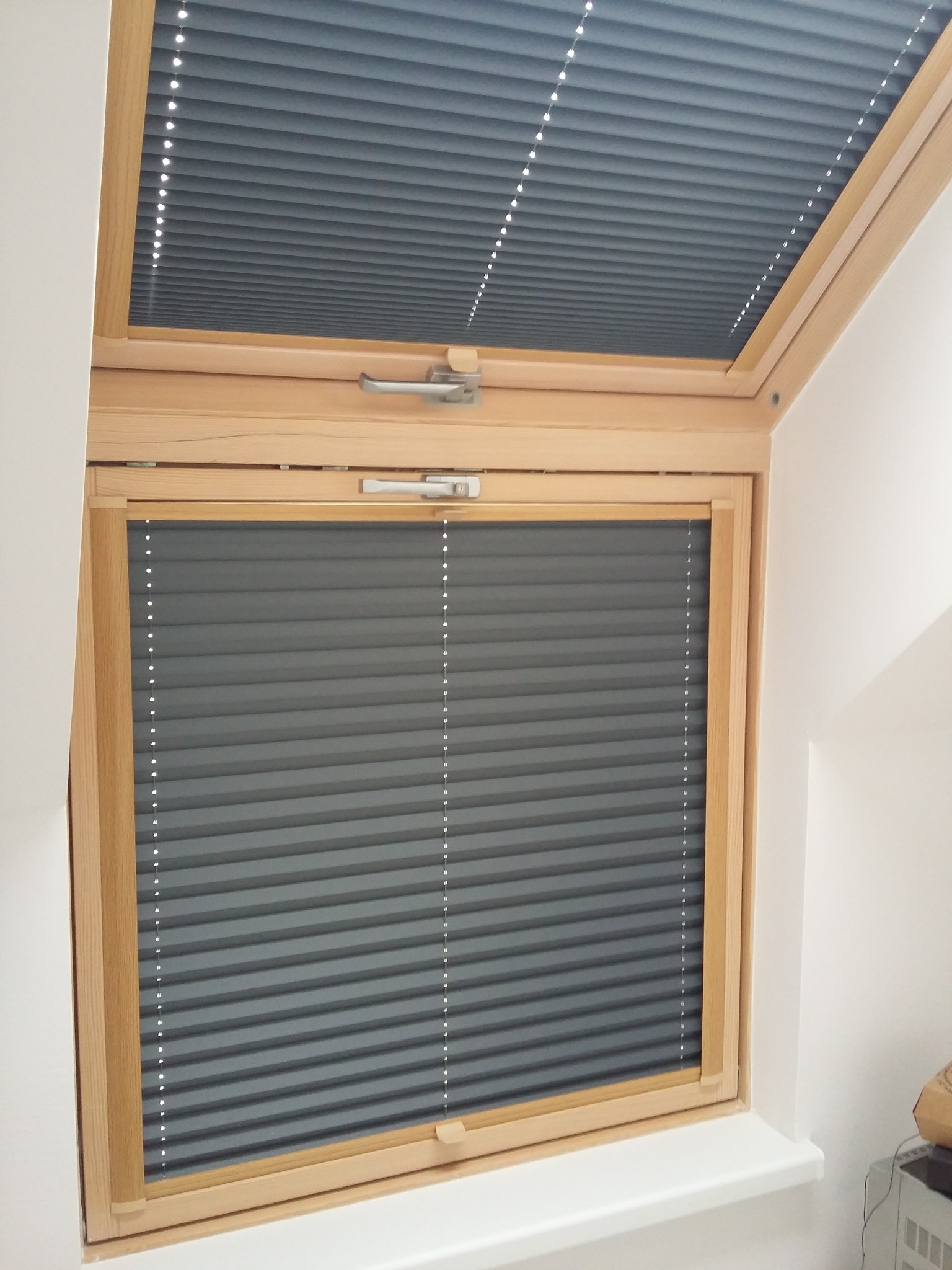 Szare, harmonijkowe osłony okienne zamontowane w drewnianej ramie okna dachowego, widoczne w dwóch częściach: górnej i dolnej.