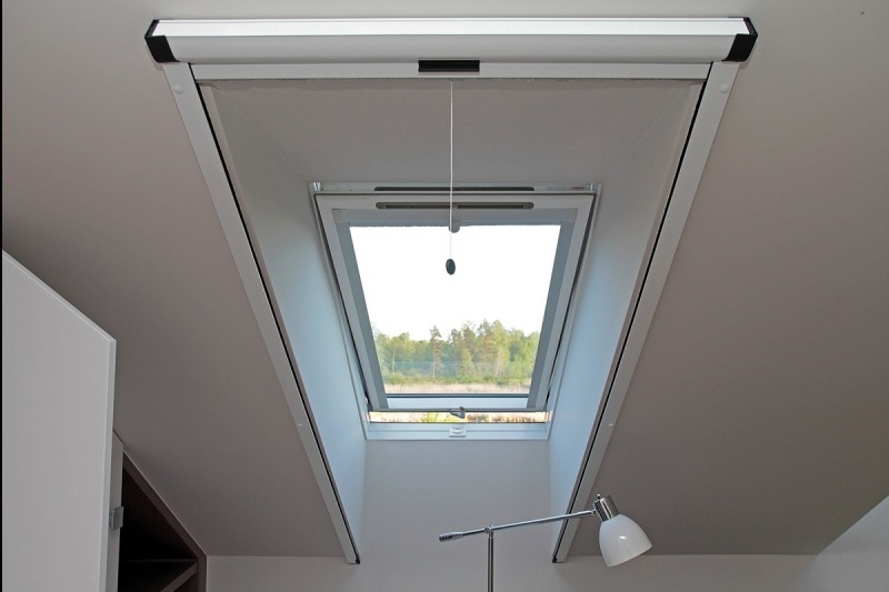 Okno dachowe z częściowo zwiniętą roletą materiałową w kasecie, widok na zewnątrz z zielenią, sznurek do regulacji zwisa pionowo.