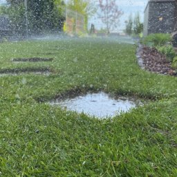 Mateusz Mamerski Usługi Ogrodnicze - Niski kąt ujęcia na świeżo nawodniony trawnik z rolki, z widoczną kałużą i kroplami wody w powietrzu, sugerujący system nawadniania w akcji.