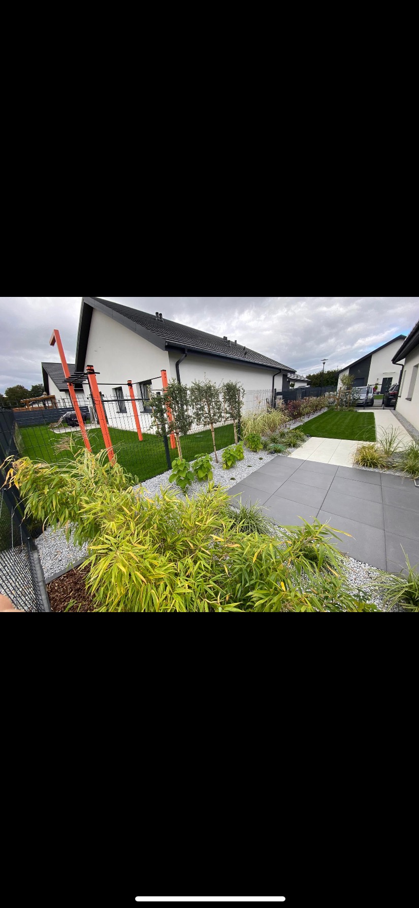 Nowoczesny ogród z szarą kostką, ozdobnym żwirem i zielenią. W tle dom z czarnym dachem i pomarańczowe elementy placu zabaw. Starannie utrzymany trawnik.