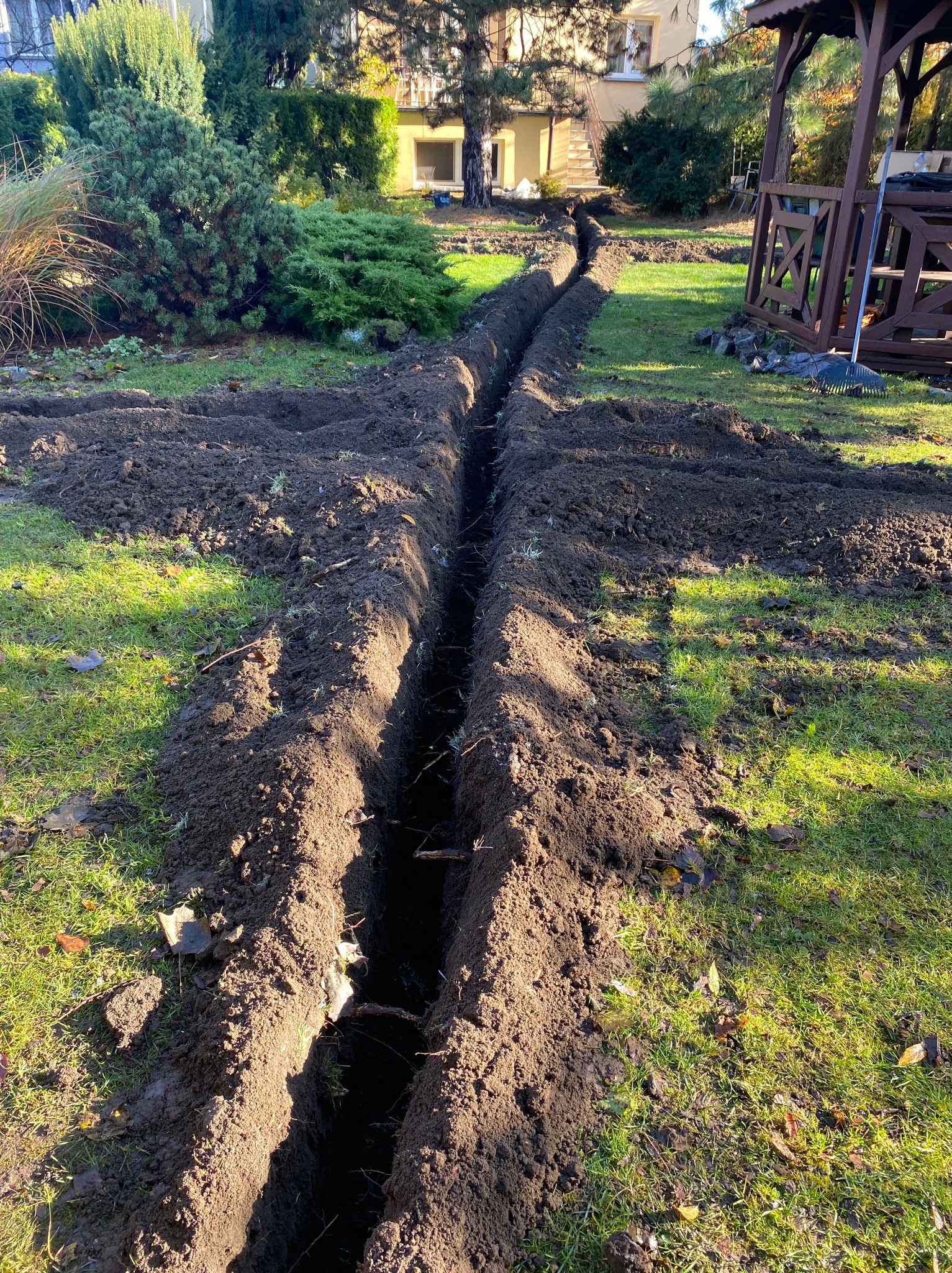 Wykopany rów w trawniku, prawdopodobnie pod system nawadniania. Widoczne ślady świeżej ziemi i fragment altany w tle. Ujęcie z perspektywy ziemi.