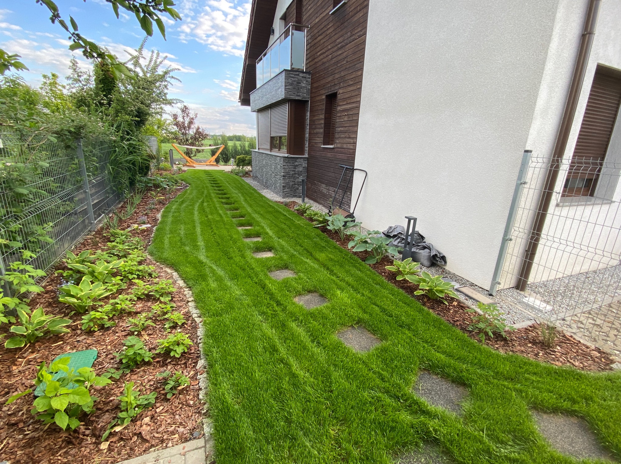 Świeżo skoszony trawnik z kamiennymi płytami prowadzącymi wzdłuż domu z drewnianą elewacją i balkonem. W tle widoczny hamak.