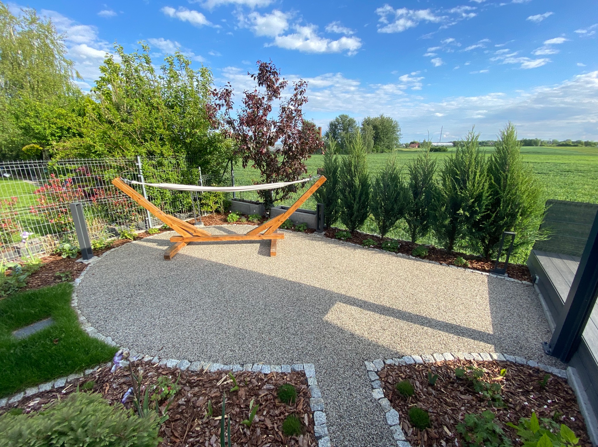 Ogród z hamakiem na żwirowym podłożu, obrzeża z kostki granitowej, rabaty z korą i roślinami, w tle zielone tuje i drzewa. Słoneczny dzień, błękitne niebo.