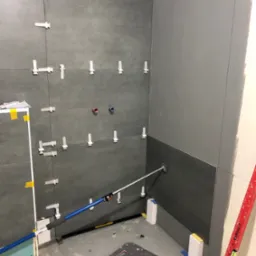 Ściana łazienki w trakcie układania szarych płytek, z użyciem krzyżyków dystansowych i poziomicy. Widoczne niebieskie rurki instalacji wodnej i narzędzie do układania płytek z przyssawką.