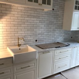 KRZYSZTOF DOKRZEWSKI - Wykończona kuchnia w stylu klasycznym: białe szafki z frezowanymi frontami i złotymi uchwytami, biała ceramiczna umywalka farmerska z mosiężną baterią, ściana wyłożona białymi, prostokątnymi...