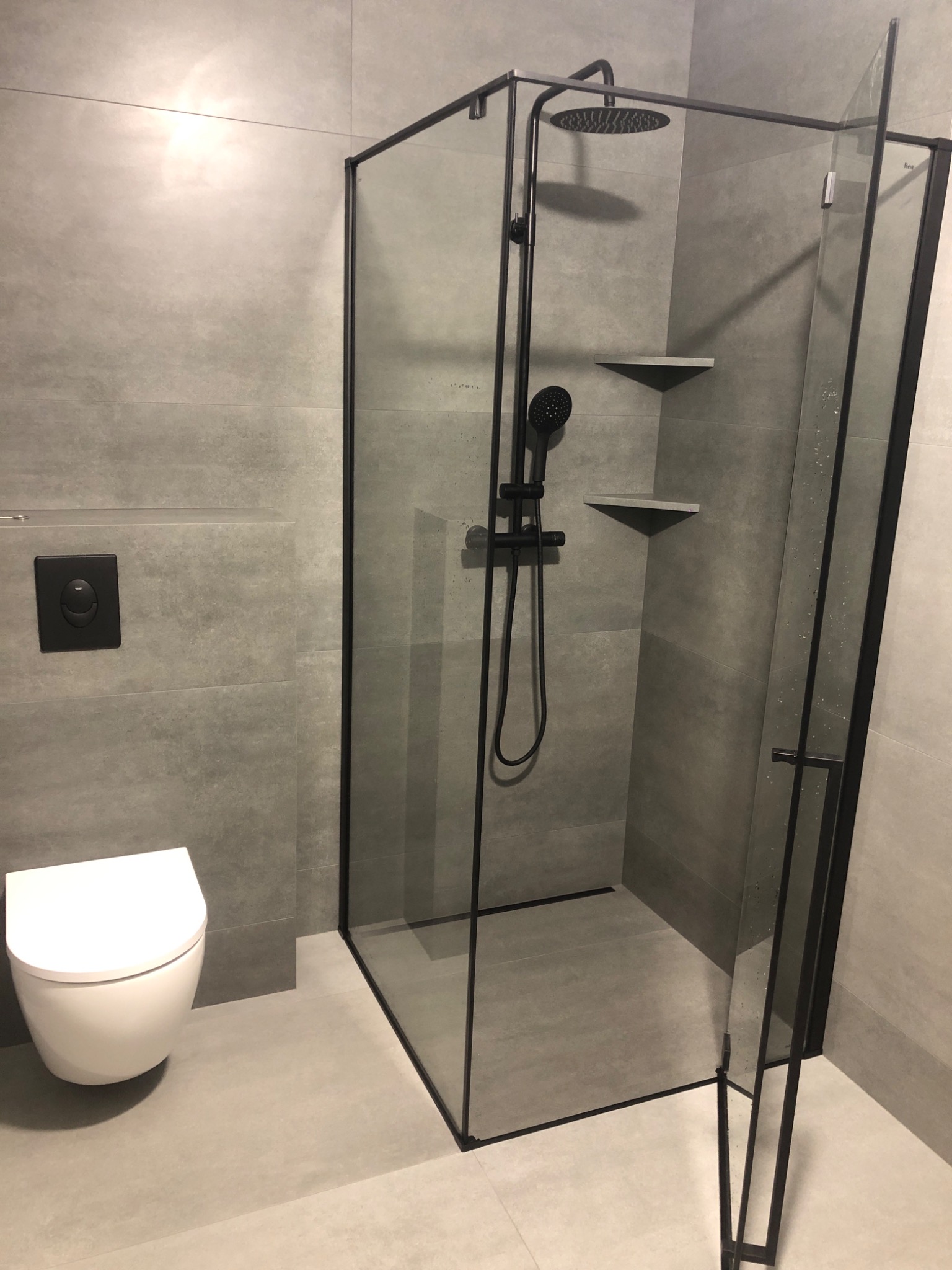 Nowoczesna łazienka z kabiną prysznicową typu walk-in z czarnymi profilami, wisząca toaleta i szara płytka wielkoformatowa.