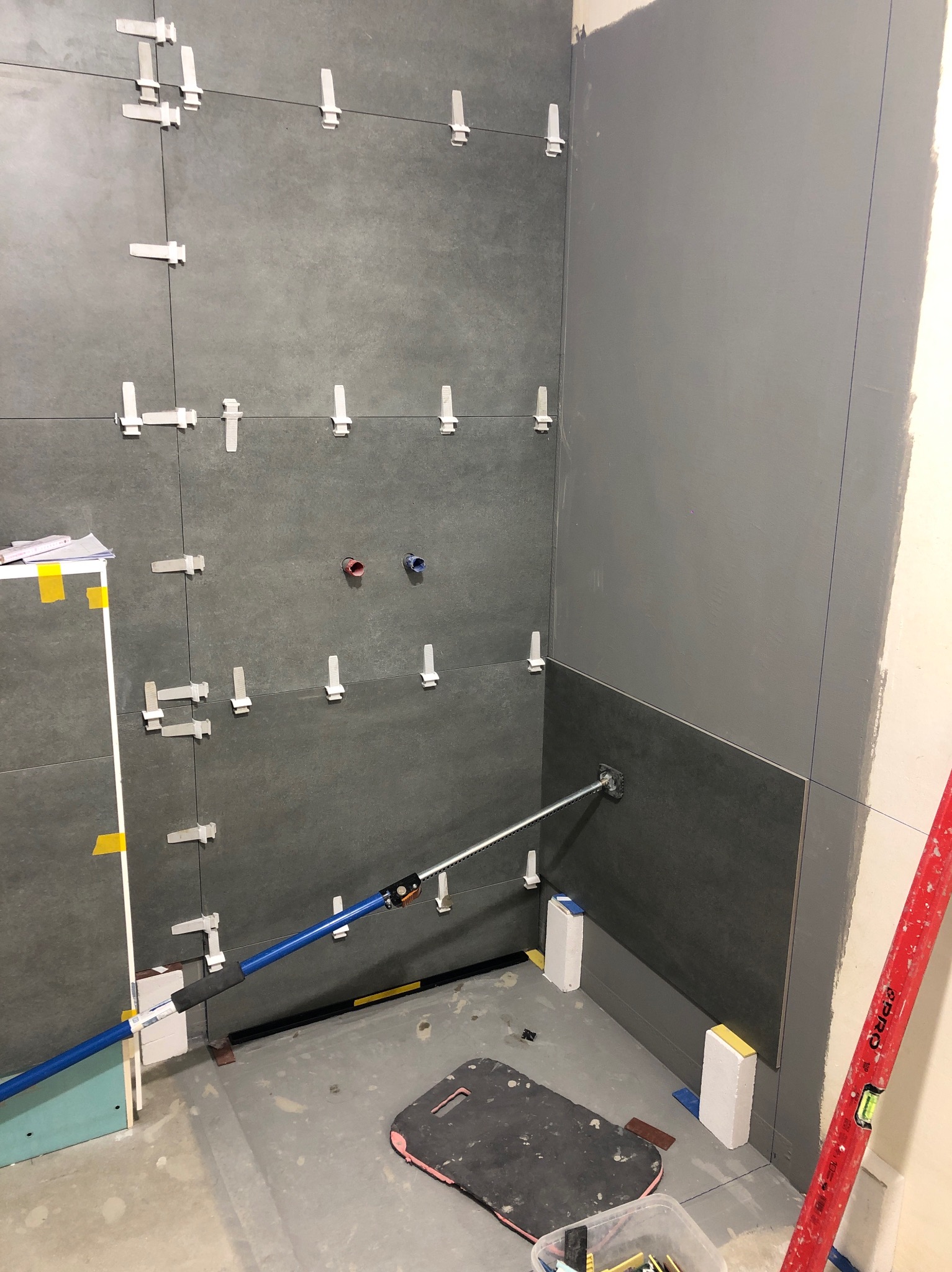 Ściana łazienki w trakcie układania szarych płytek, z użyciem krzyżyków dystansowych i poziomicy. Widoczne niebieskie rurki instalacji wodnej i narzędzie do układania płytek z przyssawką.