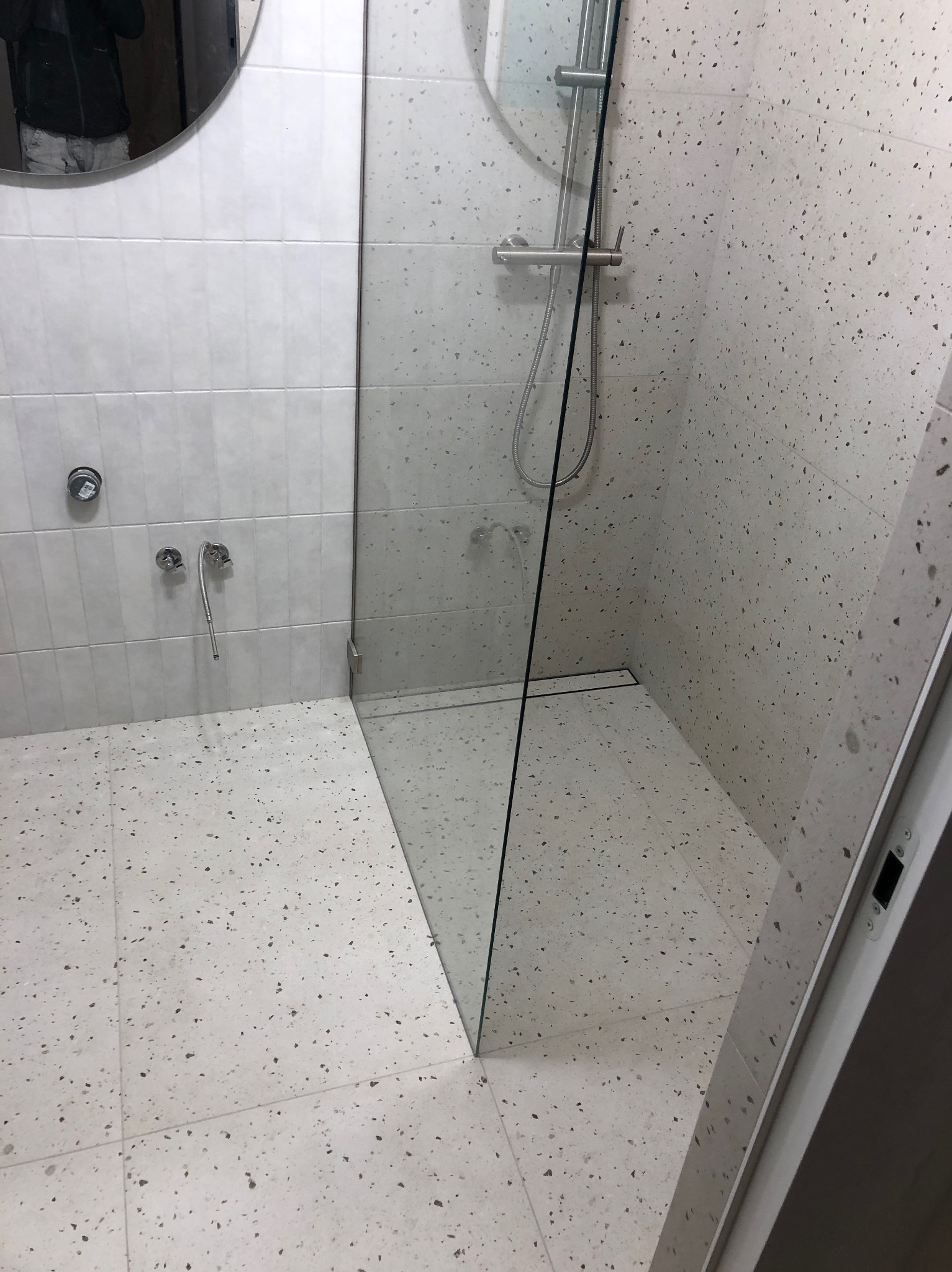 Nowoczesna kabina prysznicowa z deszczownicą, wykończona płytkami imitującymi lastryko, z minimalistycznym odpływem liniowym i lustrem w okrągłej ramie.