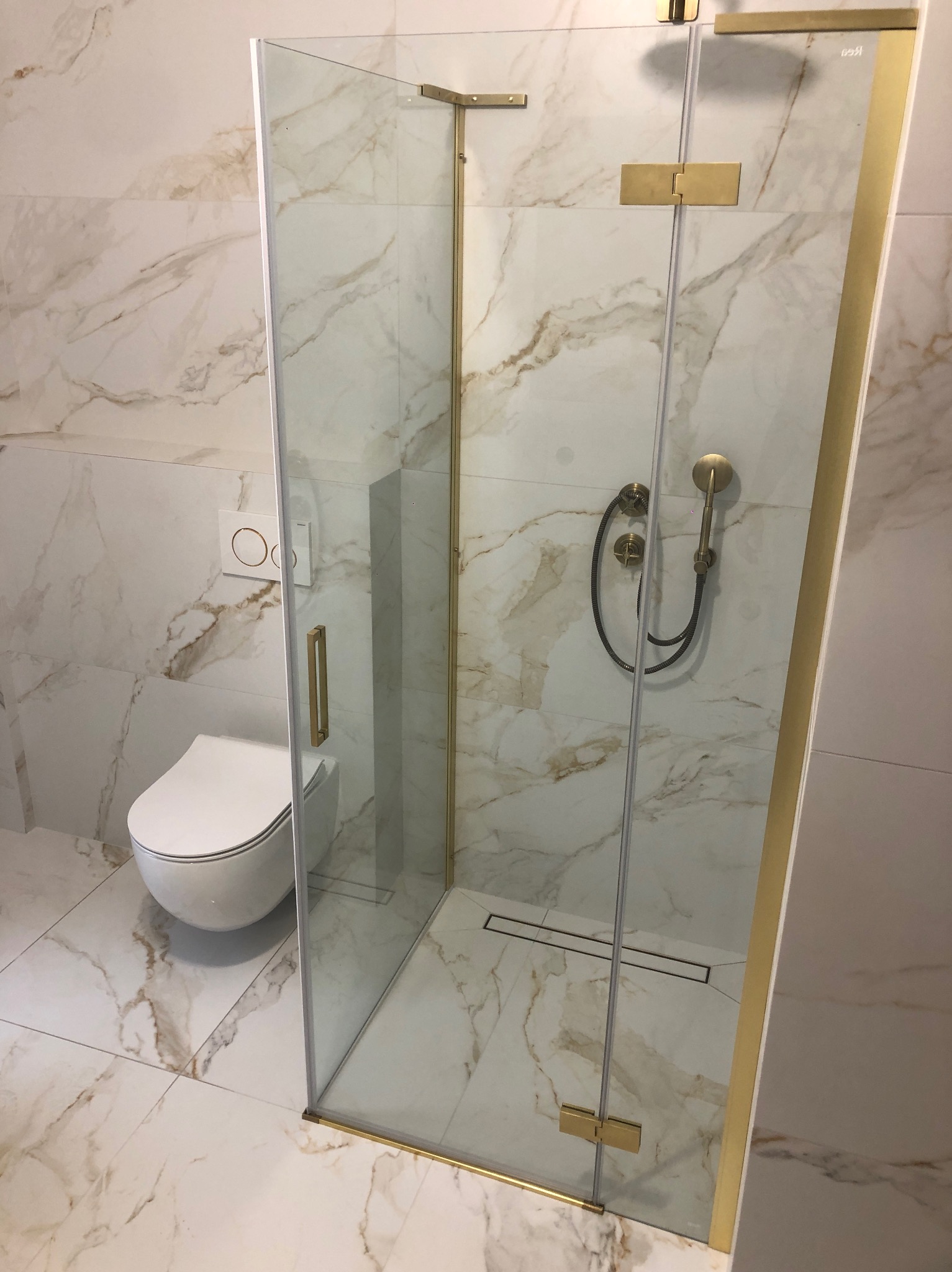 Nowoczesna łazienka z kabiną prysznicową ze złotymi okuciami i deszczownicą w stylu retro, marmurowe płytki na ścianach i podłodze, wisząca toaleta z przyciskiem spłukującym w kolorze złotym.