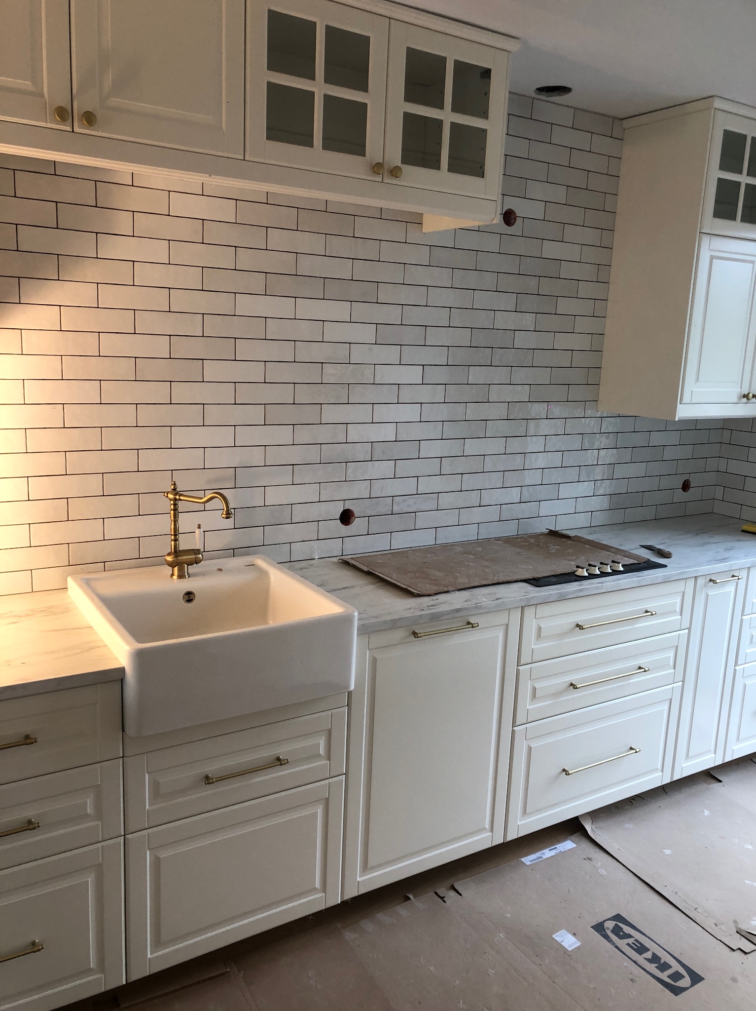 Wykończona kuchnia w stylu klasycznym: białe szafki z frezowanymi frontami i złotymi uchwytami, biała ceramiczna umywalka farmerska z mosiężną baterią, ściana wyłożona białymi, prostokątnymi...