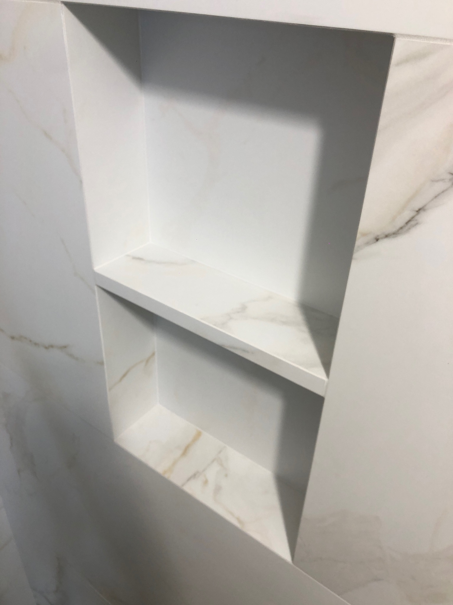 Wnęka z półkami w łazience, wykończona białymi płytkami imitującymi marmur z delikatnym żyłkowaniem w odcieniach złota.