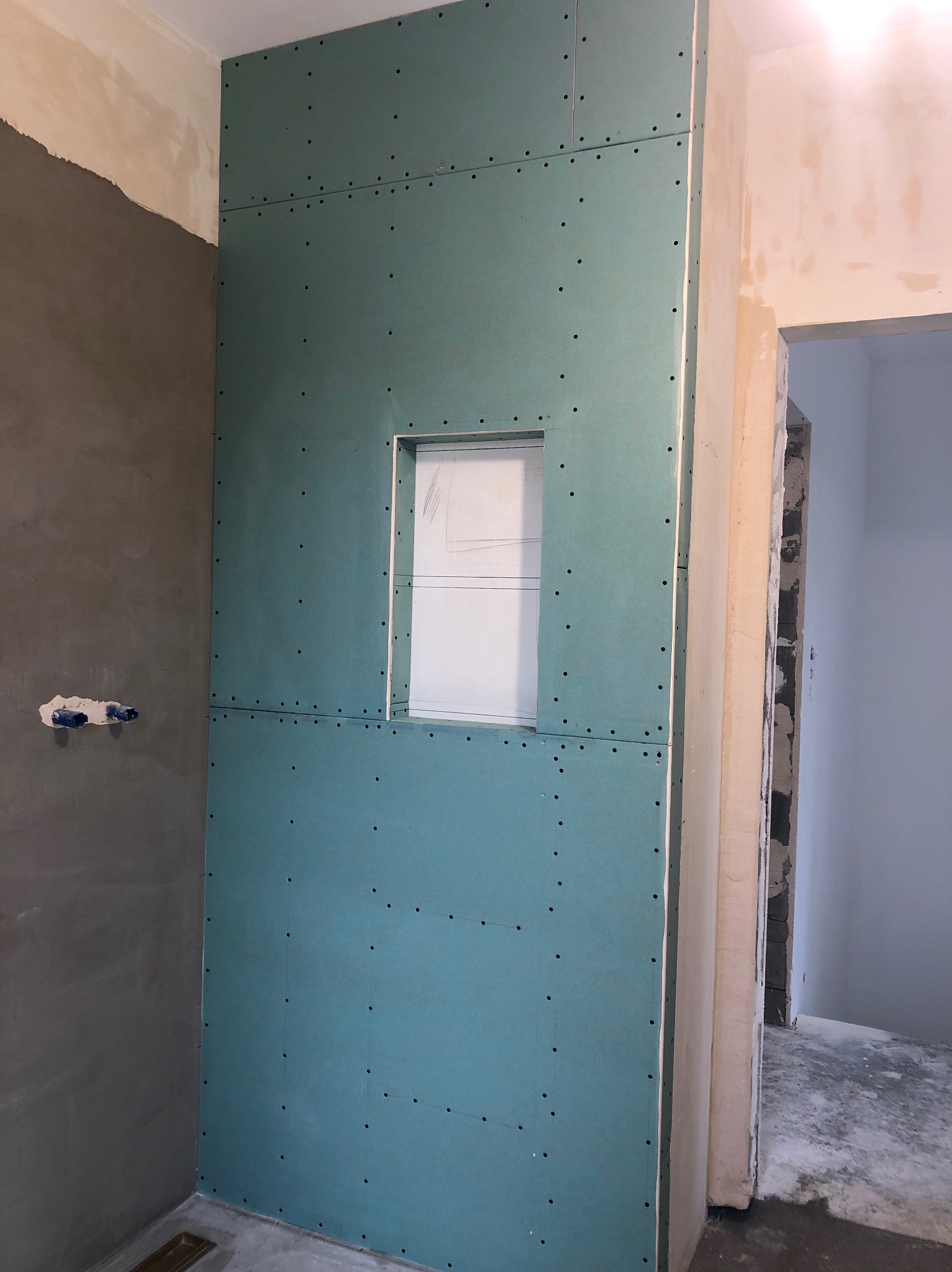 Ściana w łazience w trakcie wykańczania, pokryta zielonymi płytami gipsowo-kartonowymi z wyciętym otworem, widoczne ślady po szpachlowaniu na sąsiedniej ścianie i wystające rury instalacji wodnej.