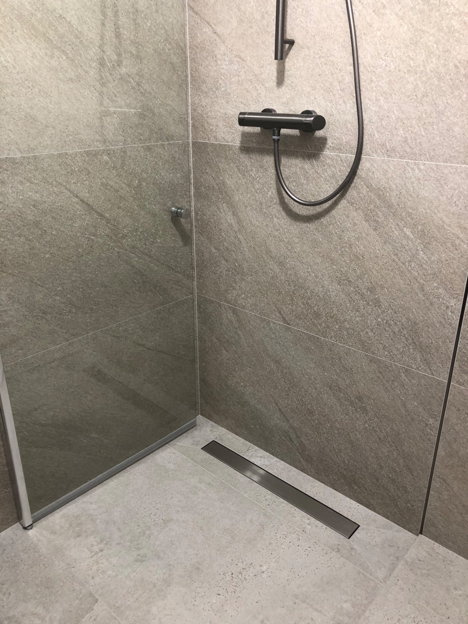 Nowoczesna kabina prysznicowa walk-in z odpływem liniowym i baterią podtynkową w odcieniach szarości, wykończona płytami imitującymi kamień.