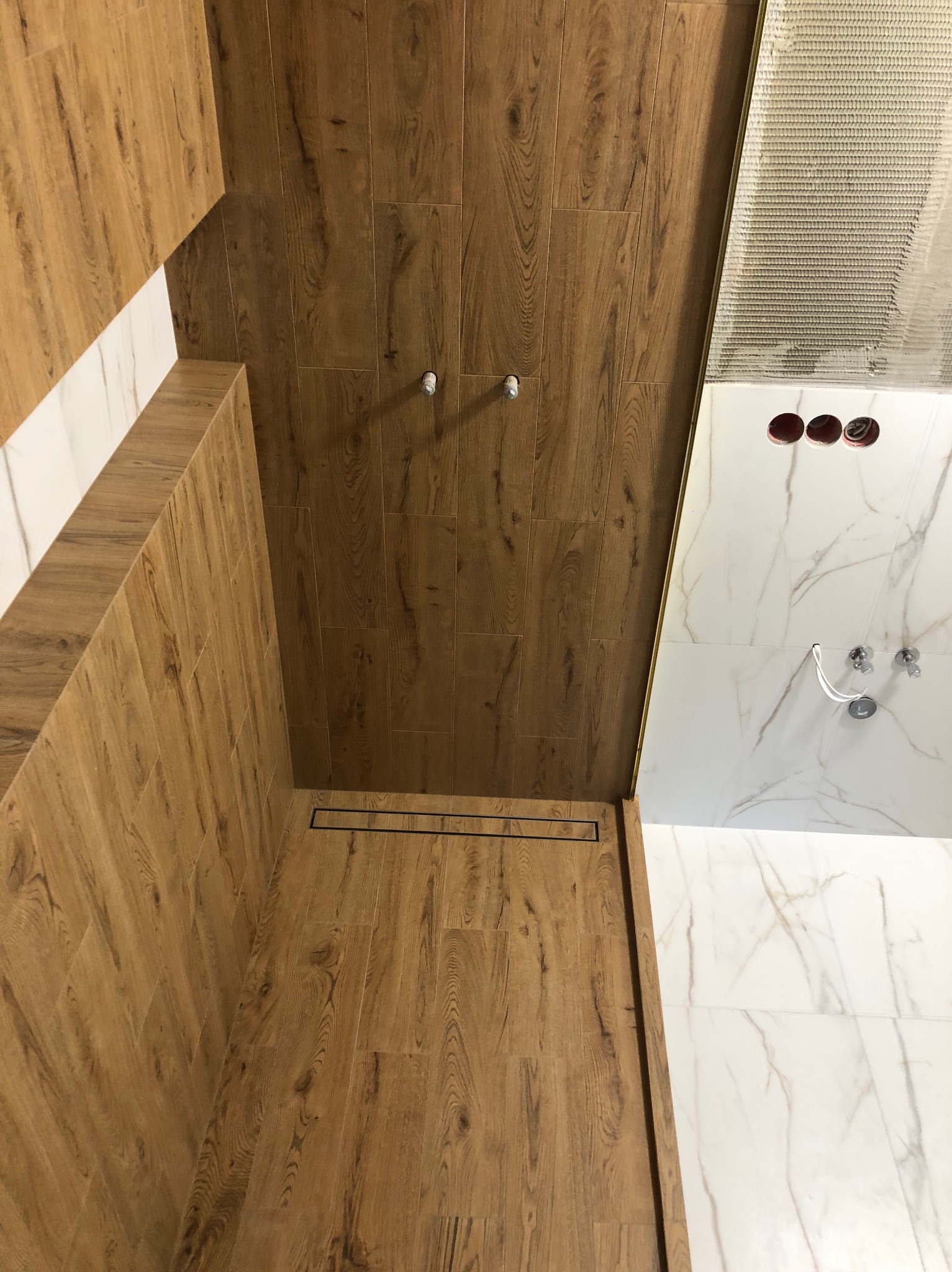 Wnętrze łazienki w trakcie wykańczania: drewnopodobne płytki na ścianach i podłodze kabiny prysznicowej z odpływem liniowym, kontrastujące z białymi marmurkowymi płytkami na ścianie obok, widoczne...