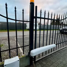 Czarna, metalowa brama z automatycznym napędem i pomarańczowym światłem ostrzegawczym, widok z bliska na mechanizm i skrzynkę sterującą przy ziemi, na zewnątrz widoczny samochód i przyczepa...