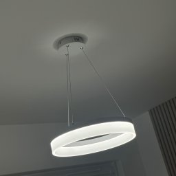 ANVO Patryk Andryszewski - Nowoczesna, okrągła lampa LED wisząca z białym kloszem i cienkimi linkami mocującymi, zamontowana na suficie w pomieszczeniu z panelami dekoracyjnymi na ścianie.