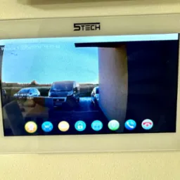 Ekran wideodomofonu marki STECH zamontowany na ścianie, wyświetlający obraz z kamery na podjeździe z zaparkowanymi samochodami i komunikatem 'Brak karty'.