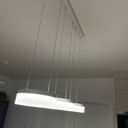 ANVO Patryk Andryszewski - Nowoczesna, podłużna lampa LED z trzema elementami świetlnymi zawieszona na cienkich linkach, zamontowana na białym suficie w pomieszczeniu z białymi drzwiami i fragmentem zabudowy kuchennej.