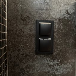 ANVO Patryk Andryszewski - Dwa czarne, kwadratowe włączniki światła z oznaczeniem IP44, zamontowane na ciemnej, teksturowanej ścianie obok fragmentu ściany wyłożonej czarną mozaiką.