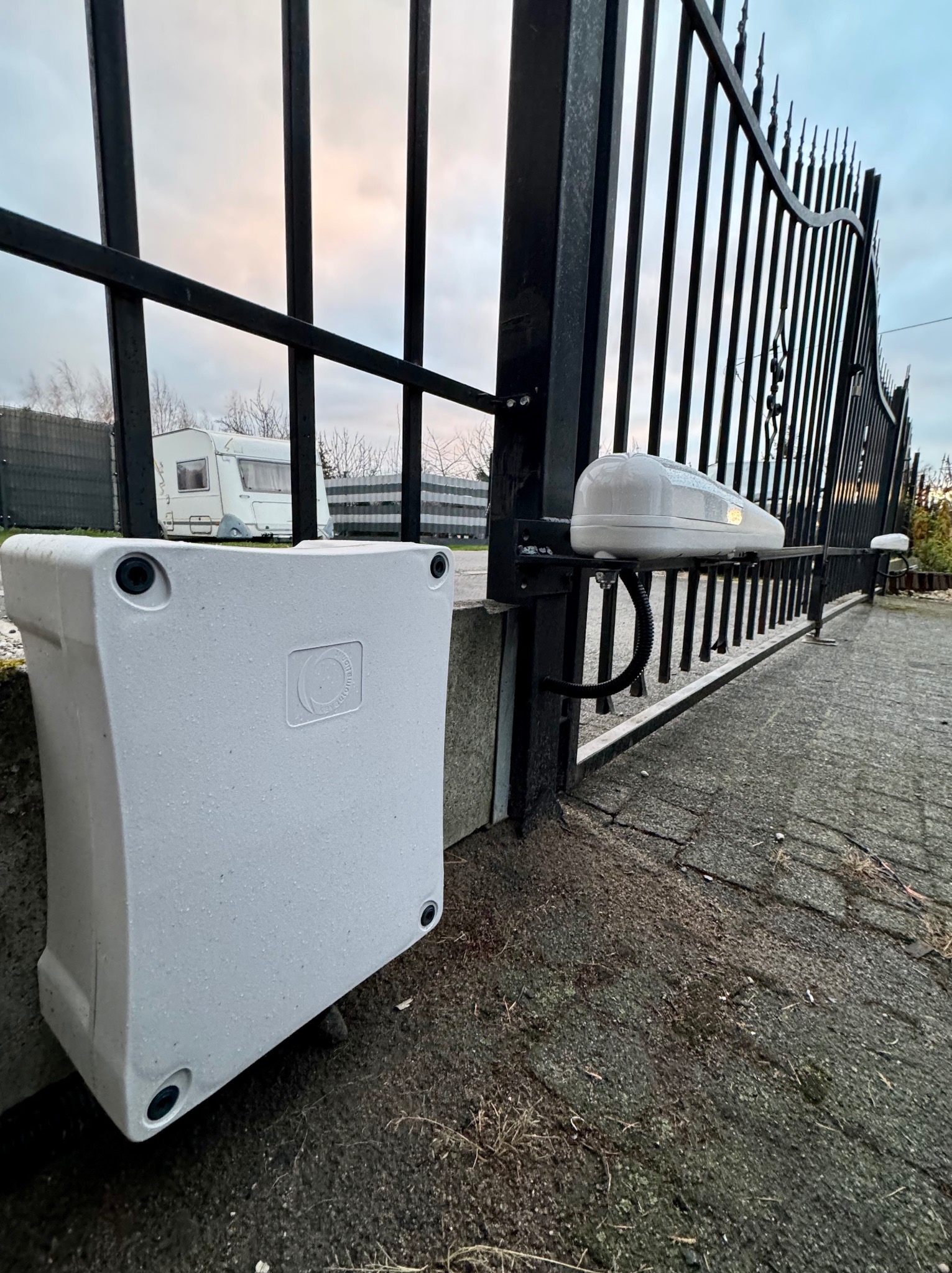 Biała obudowa sterownika i napęd bramy wjazdowej z czarnego metalu, widok na szary bruk i przyczepę kempingową w tle.