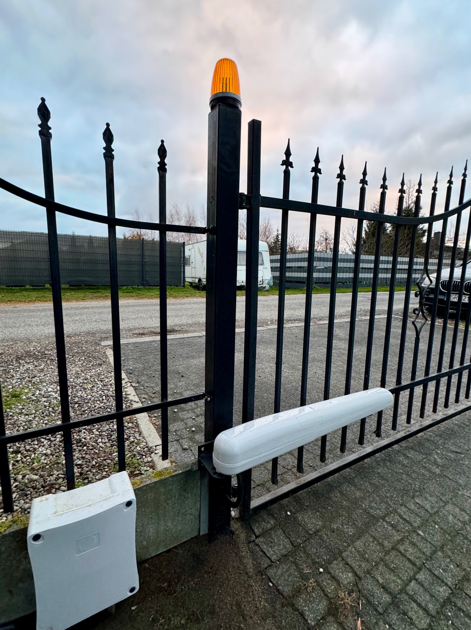 Czarna, metalowa brama z automatycznym napędem i pomarańczowym światłem ostrzegawczym, widok z bliska na mechanizm i skrzynkę sterującą przy ziemi, na zewnątrz widoczny samochód i przyczepa...