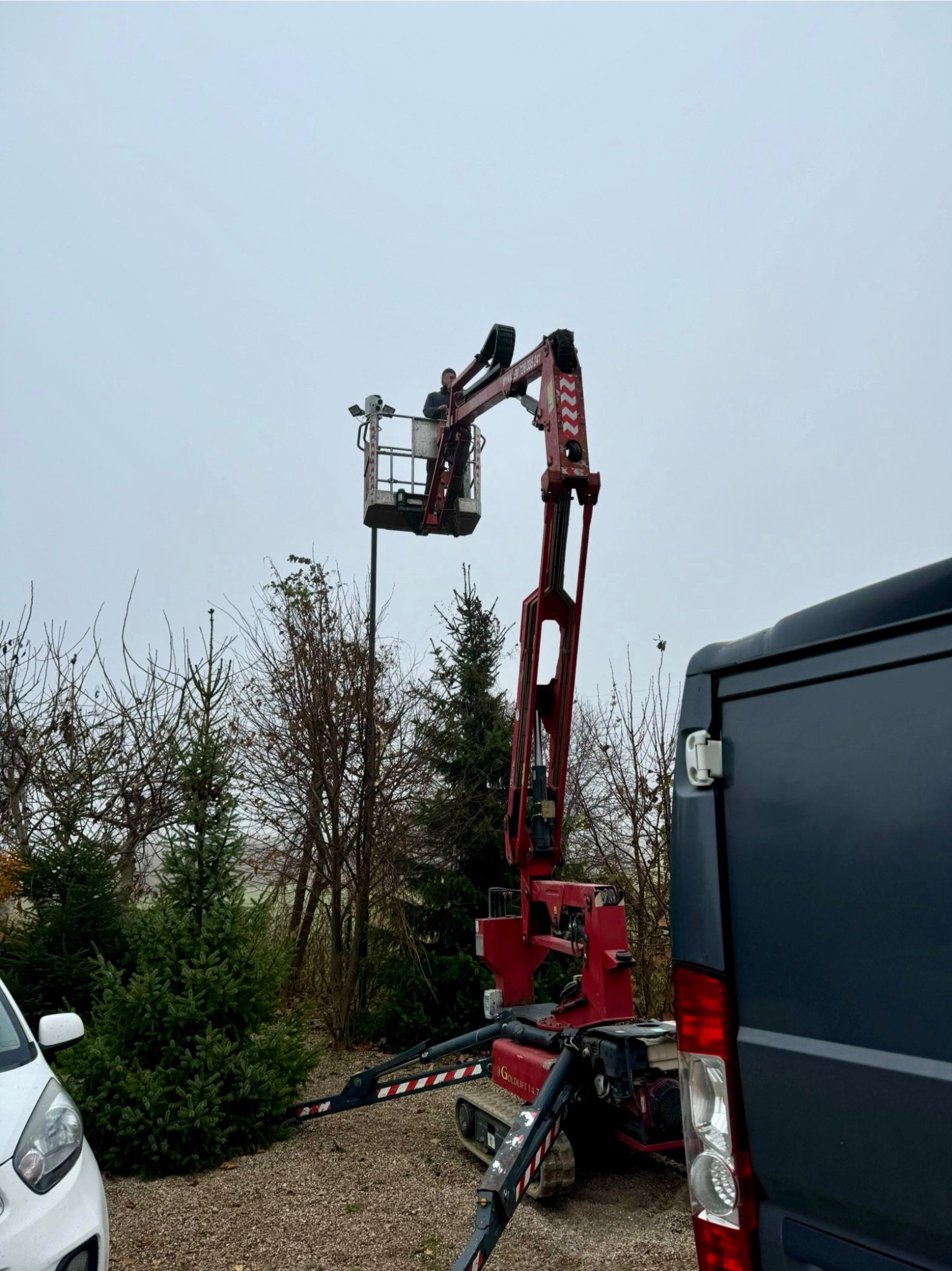 Czerwony podnośnik koszowy na gąsienicach, z pracownikiem w koszu, ustawiony na żwirowej powierzchni obok zaparkowanego busa i samochodu osobowego, na tle drzew i pochmurnego nieba. Prace...