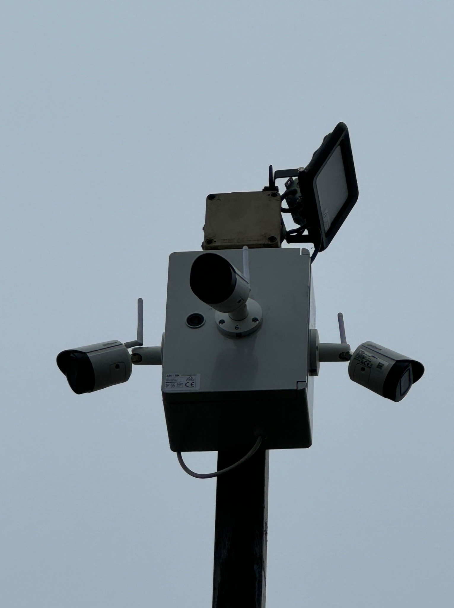 Zestaw kamer monitoringu zewnętrznego z reflektorem, zamontowany na słupie, widok z dołu na szare niebo.