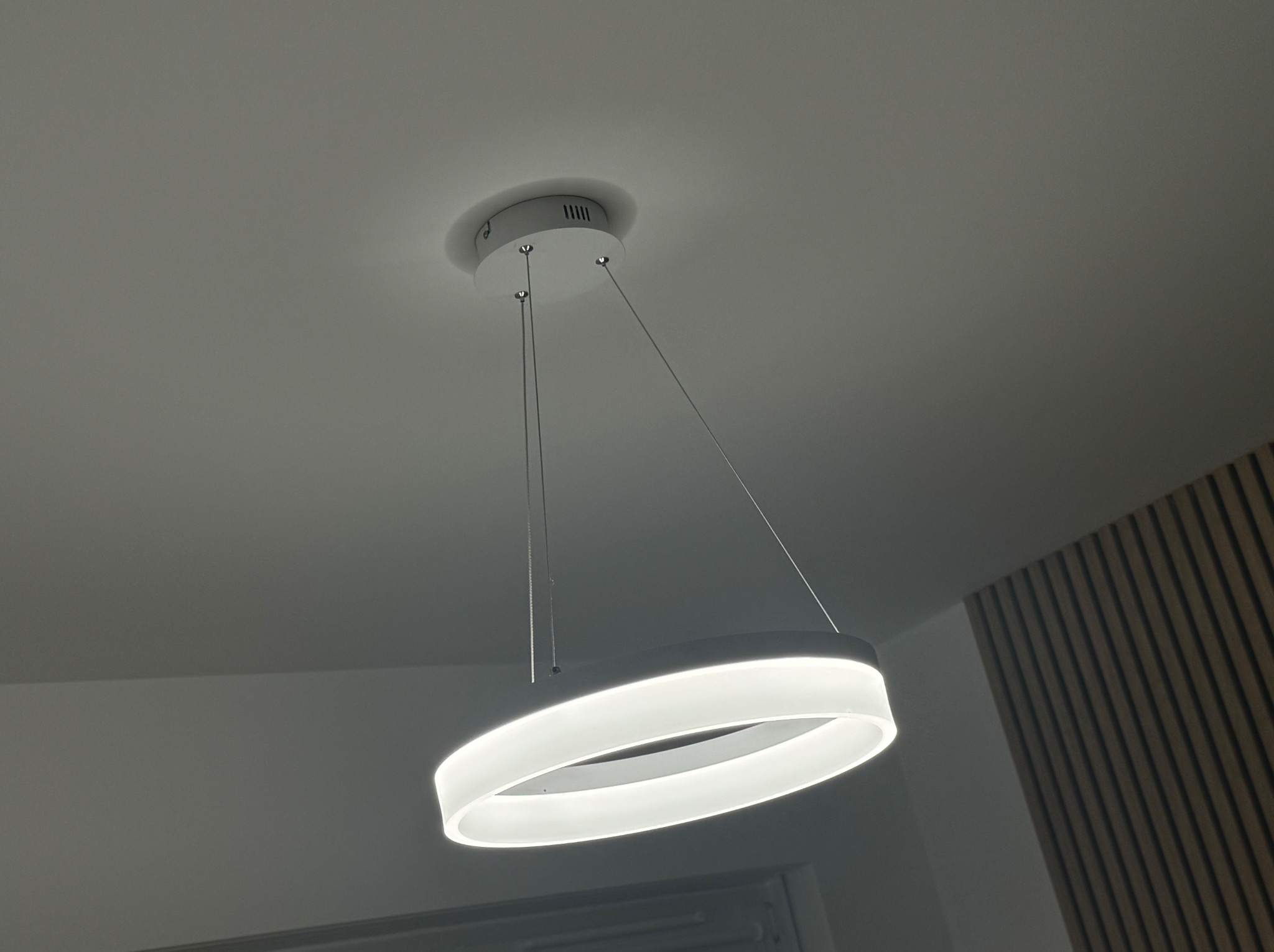 Nowoczesna, okrągła lampa LED wisząca z białym kloszem i cienkimi linkami mocującymi, zamontowana na suficie w pomieszczeniu z panelami dekoracyjnymi na ścianie.