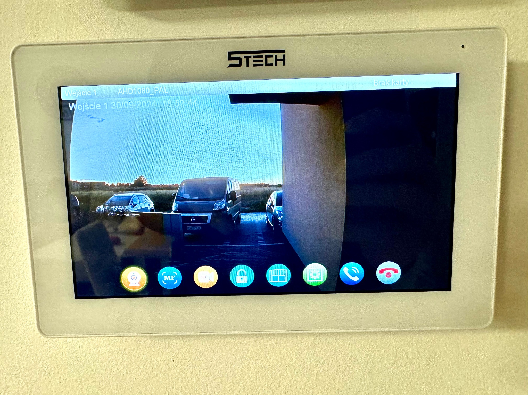 Ekran wideodomofonu marki STECH zamontowany na ścianie, wyświetlający obraz z kamery na podjeździe z zaparkowanymi samochodami i komunikatem 'Brak karty'.