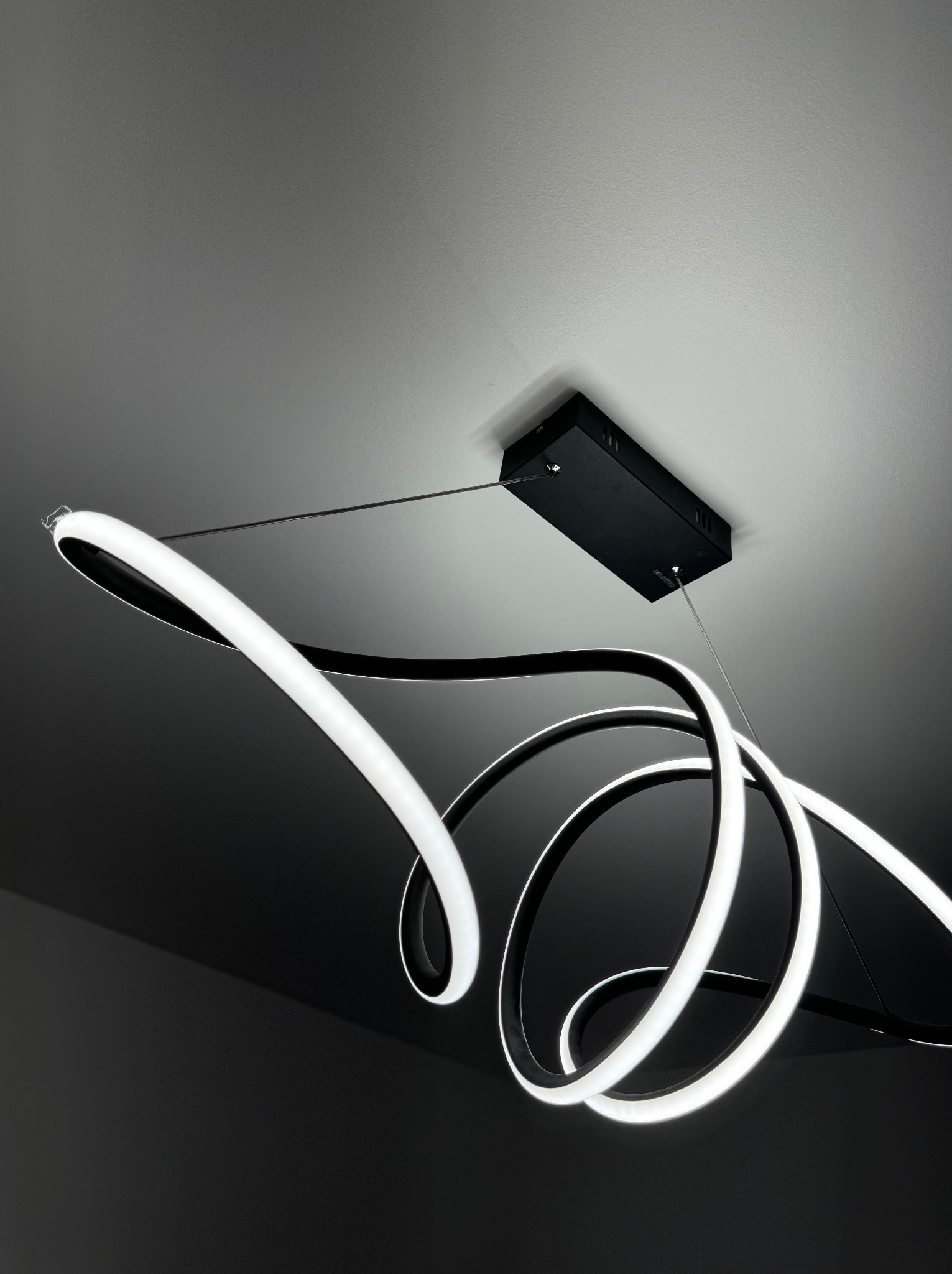 Nowoczesna lampa wisząca LED w kształcie spiralnych pierścieni, zawieszona na cienkich linkach, z czarną podsufitką, oświetlająca ciemne wnętrze.