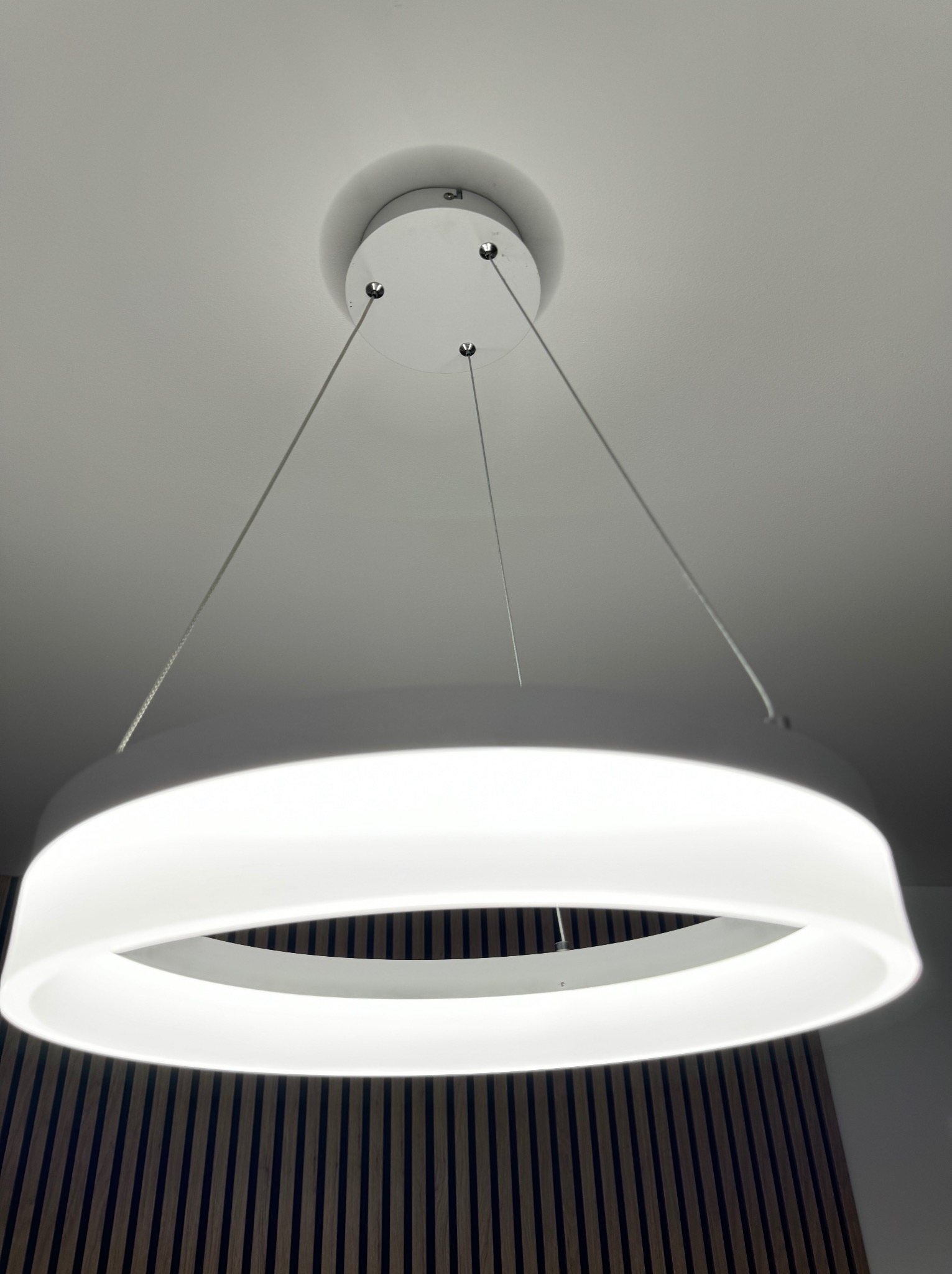 Nowoczesna lampa wisząca LED w kształcie pierścienia, zawieszona na cienkich linkach, oświetlająca ścianę z drewnianych paneli.