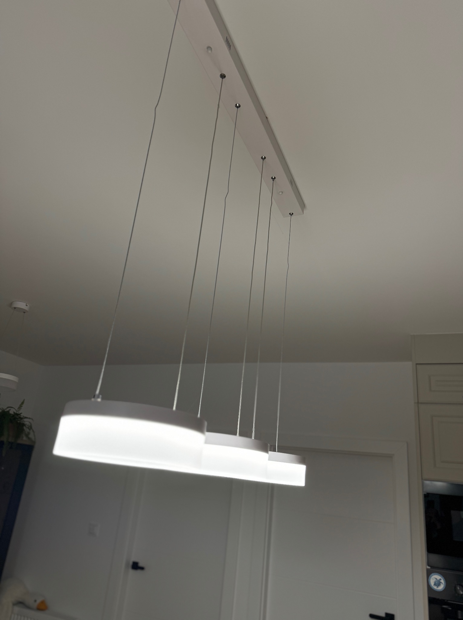 Nowoczesna, podłużna lampa LED z trzema elementami świetlnymi zawieszona na cienkich linkach, zamontowana na białym suficie w pomieszczeniu z białymi drzwiami i fragmentem zabudowy kuchennej.