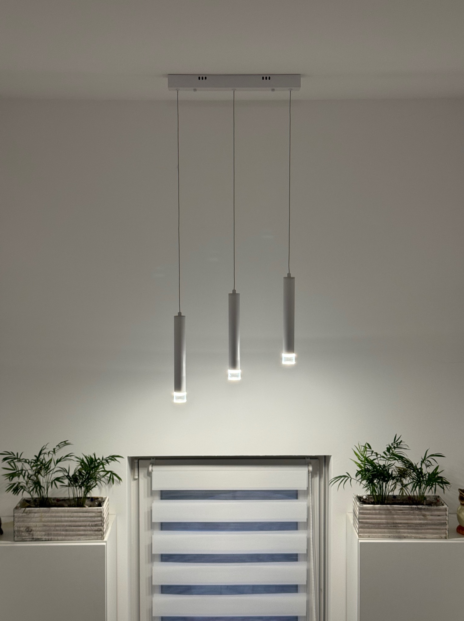 Nowoczesna lampa wisząca z trzema cylindrycznymi kloszami, oświetlająca okno z roletami dzień-noc, z doniczkami z roślinami po bokach, na tle białej ściany.