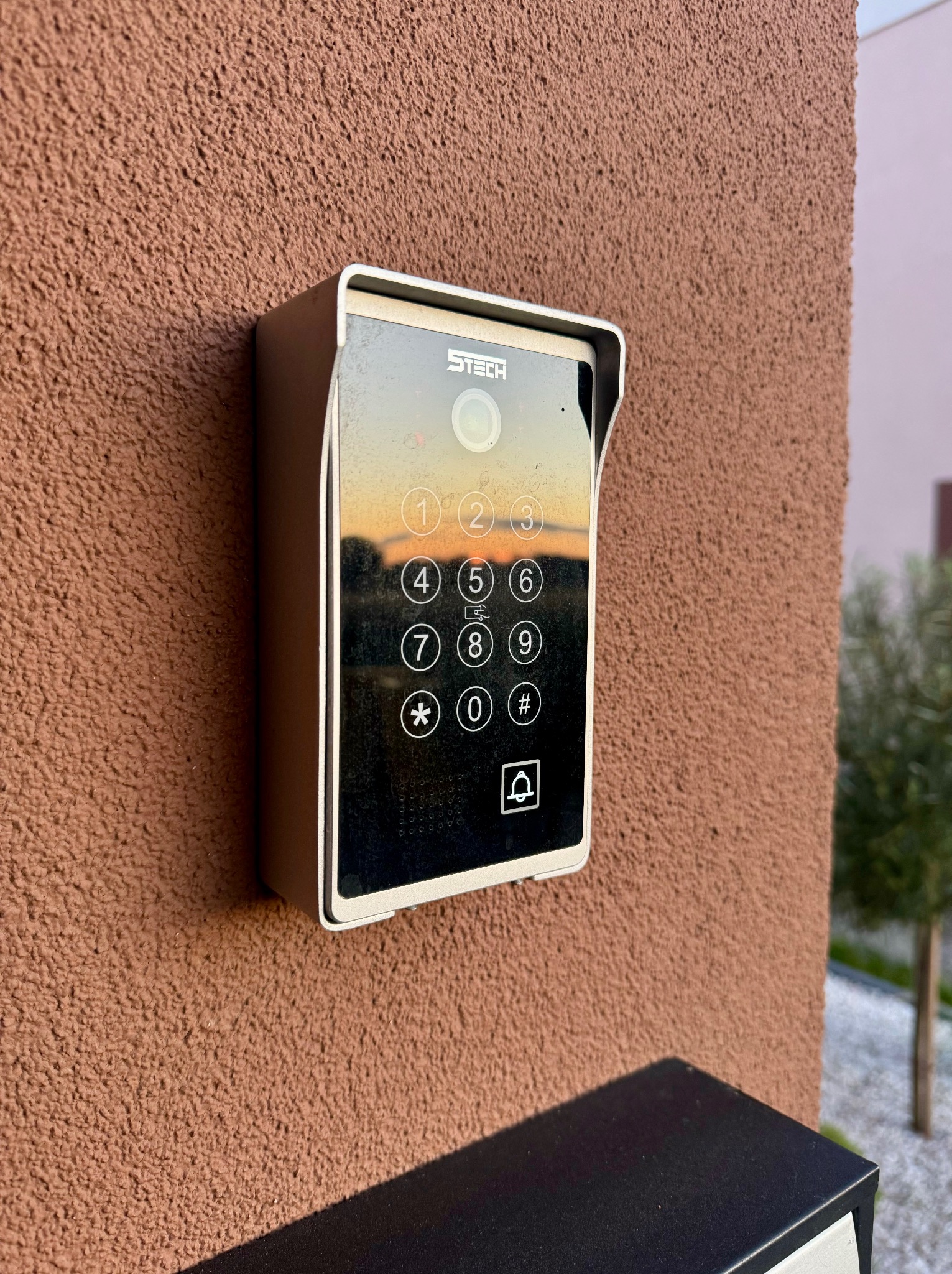 Nowoczesny wideofon z klawiaturą numeryczną marki STECH, zamontowany na brązowej elewacji budynku. W ekranie odbija się zachód słońca.