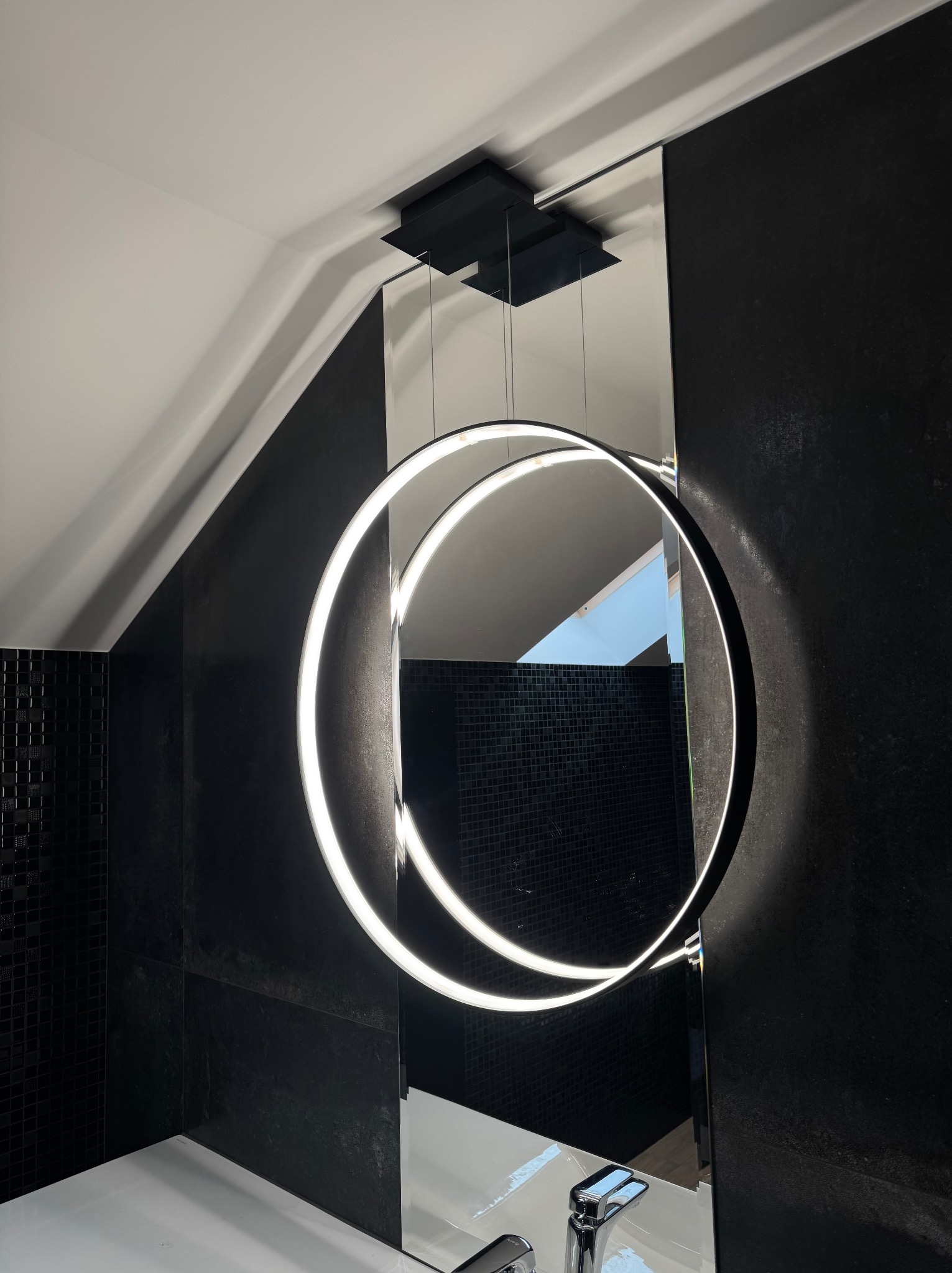 Nowoczesne oświetlenie łazienkowe: okrągłe lustro z podświetleniem LED, czarne płytki mozaikowe i minimalistyczna armatura. Stylowe wykończenie wnętrza.