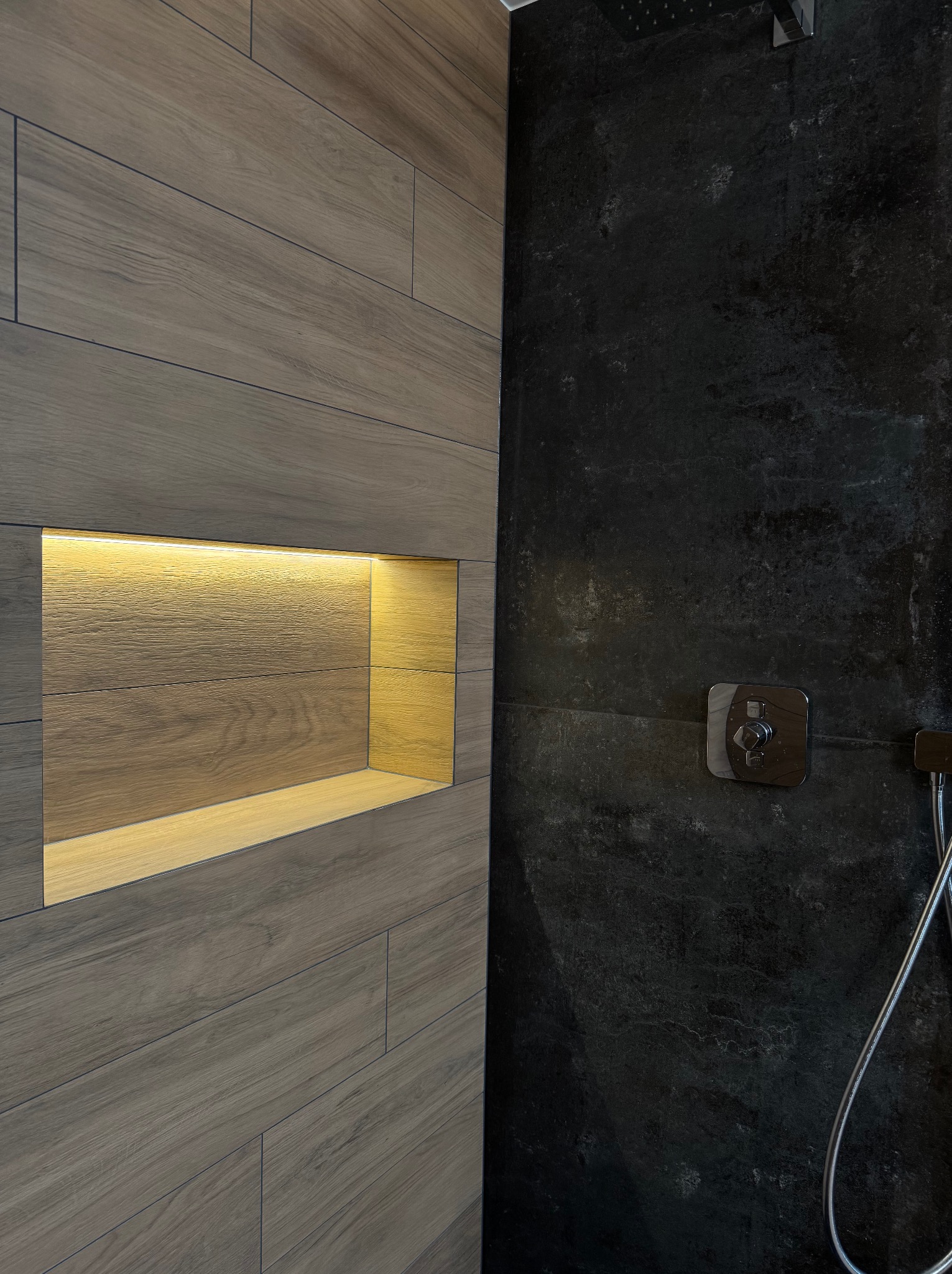 Nowoczesna kabina prysznicowa z drewnopodobnymi płytkami i wnęką z podświetleniem LED. Ciemna ściana z baterią i słuchawką prysznicową.