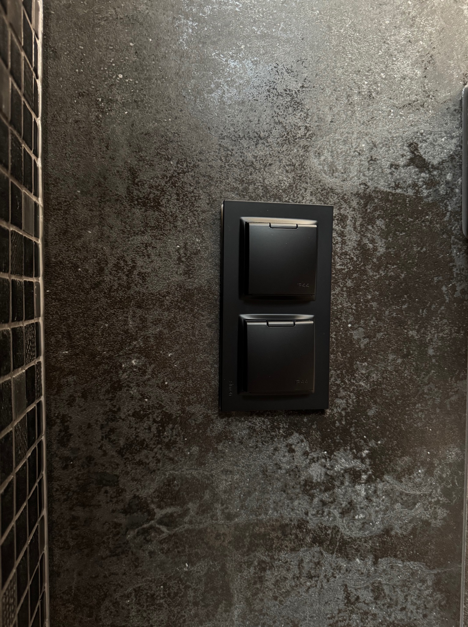 Dwa czarne, kwadratowe włączniki światła z oznaczeniem IP44, zamontowane na ciemnej, teksturowanej ścianie obok fragmentu ściany wyłożonej czarną mozaiką.