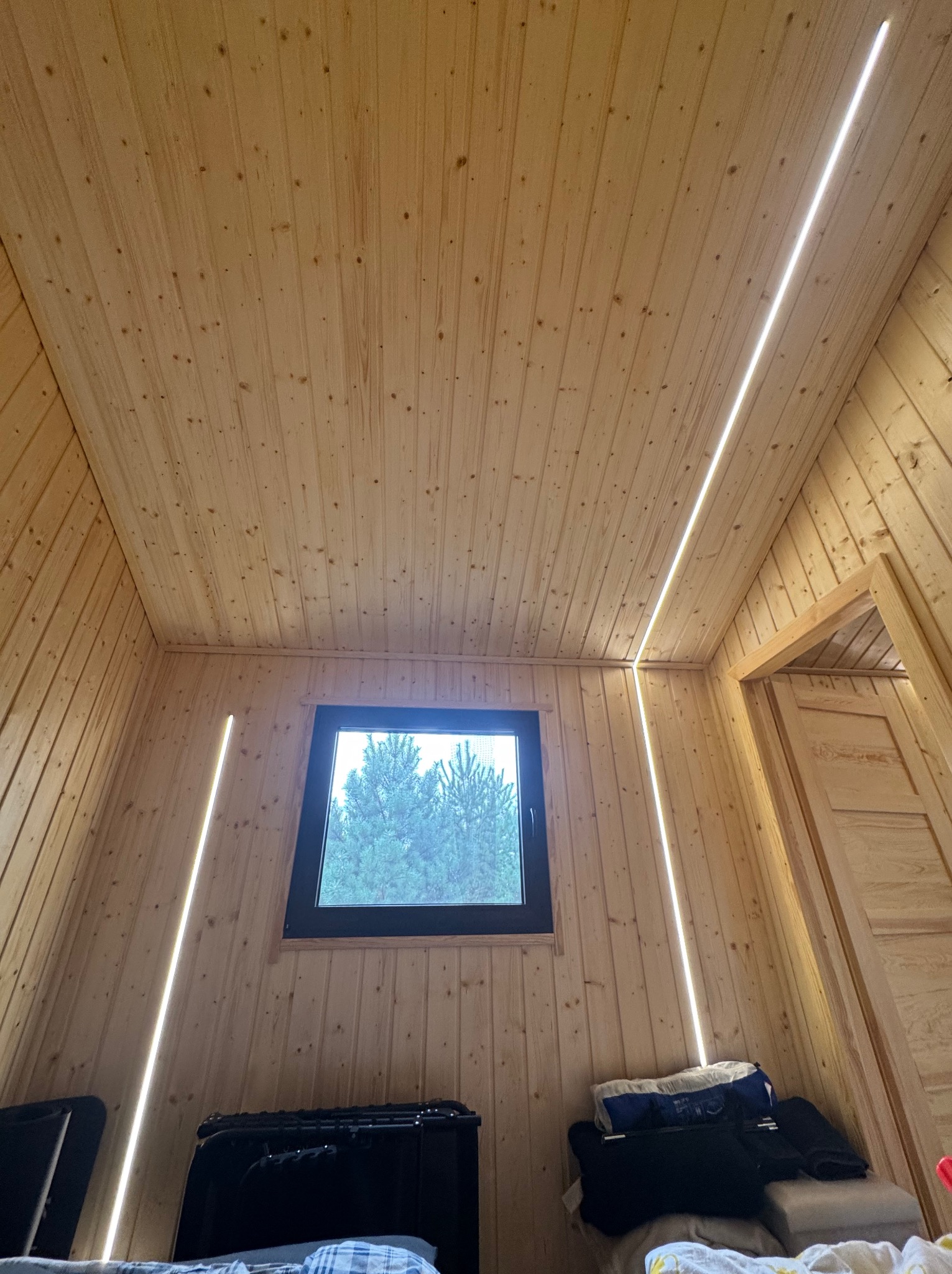 Wnętrze pokoju wykończonego boazerią z oknem i nowoczesnym oświetleniem LED w postaci pionowych linii świetlnych na ścianach i suficie. Widok na zewnątrz przez okno.