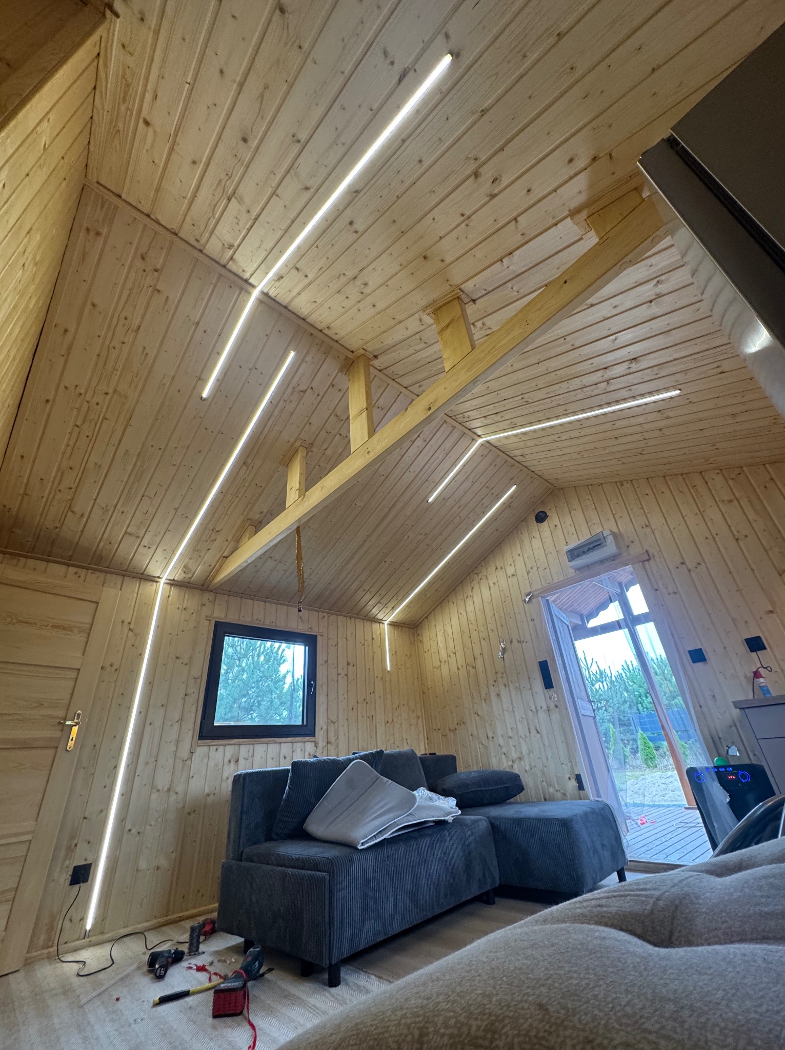 Drewniane wnętrze ze skośnym sufitem oświetlonym liniowymi lampami LED. Widoczny fragment sofy i narzędzia na podłodze sugerujące prace wykończeniowe.