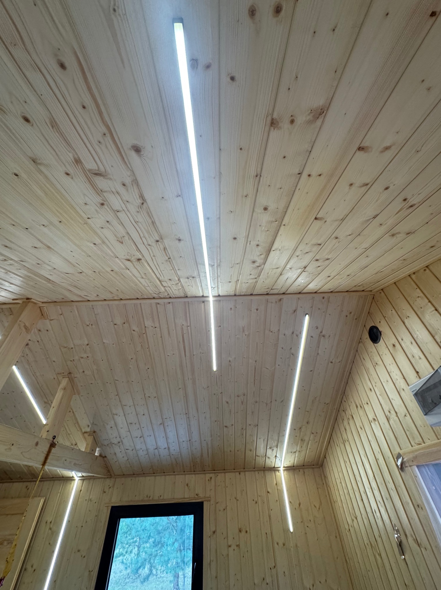 Wnętrze wykończone boazerią z zamontowanym, nowoczesnym oświetleniem LED w postaci długich, smukłych lamp. Widok na sufit i fragment ściany z oknem.