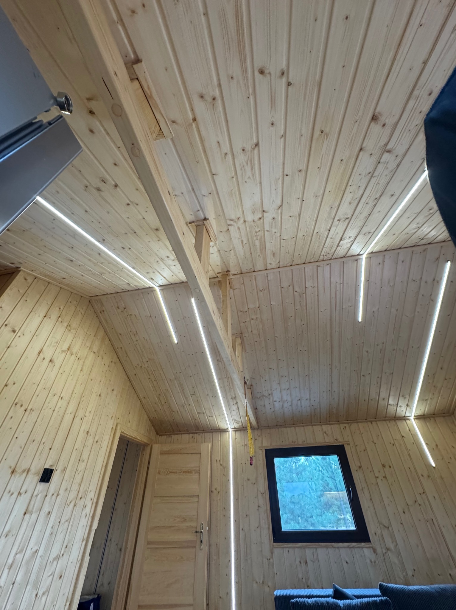 Drewniane wnętrze ze skośnym sufitem i ścianami, oświetlone pionowymi paskami LED. Widoczne okno i fragment mebla. Nietypowe oświetlenie w drewnianym domu.