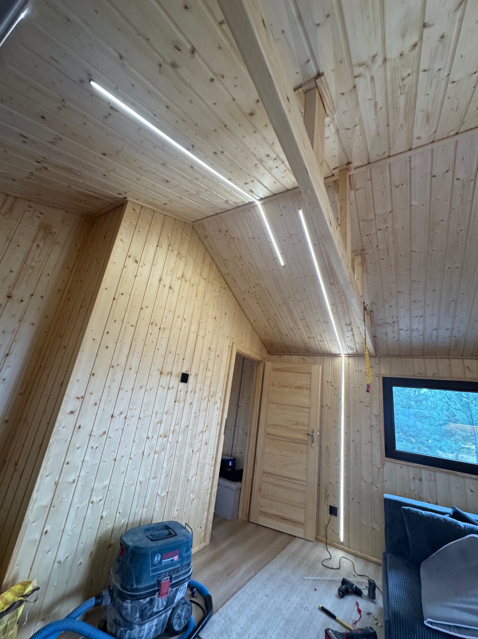 Wnętrze domu z drewnianą boazerią i zamontowanym oświetleniem LED. Widoczny odkurzacz przemysłowy i narzędzia, sugerujące prace wykończeniowe.