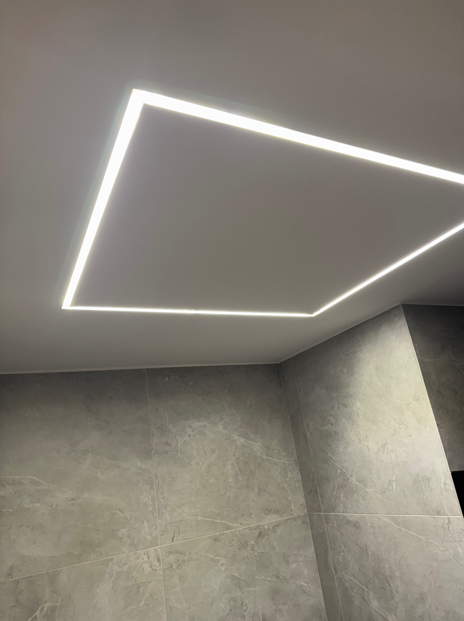 Nowoczesne oświetlenie sufitowe LED w kształcie kwadratu, wkomponowane w szarą łazienkę z marmurowymi płytkami. Minimalistyczny design, jasne, równomierne światło.