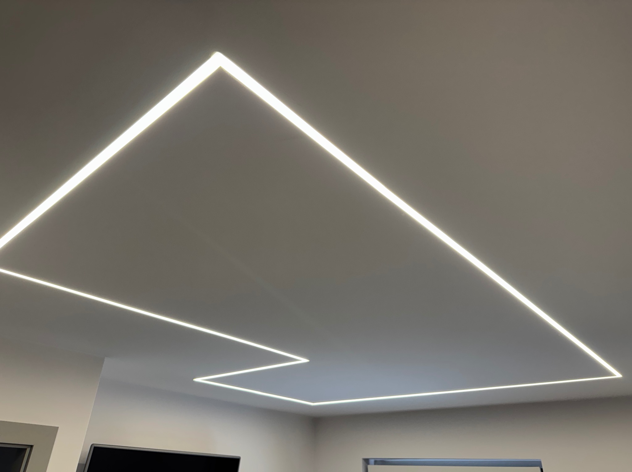 Nowoczesne oświetlenie sufitowe LED w kształcie geometrycznym, tworzące unikalny wzór świetlny w jasnym pomieszczeniu. Minimalistyczny design z precyzyjnie ułożonymi liniami światła.
