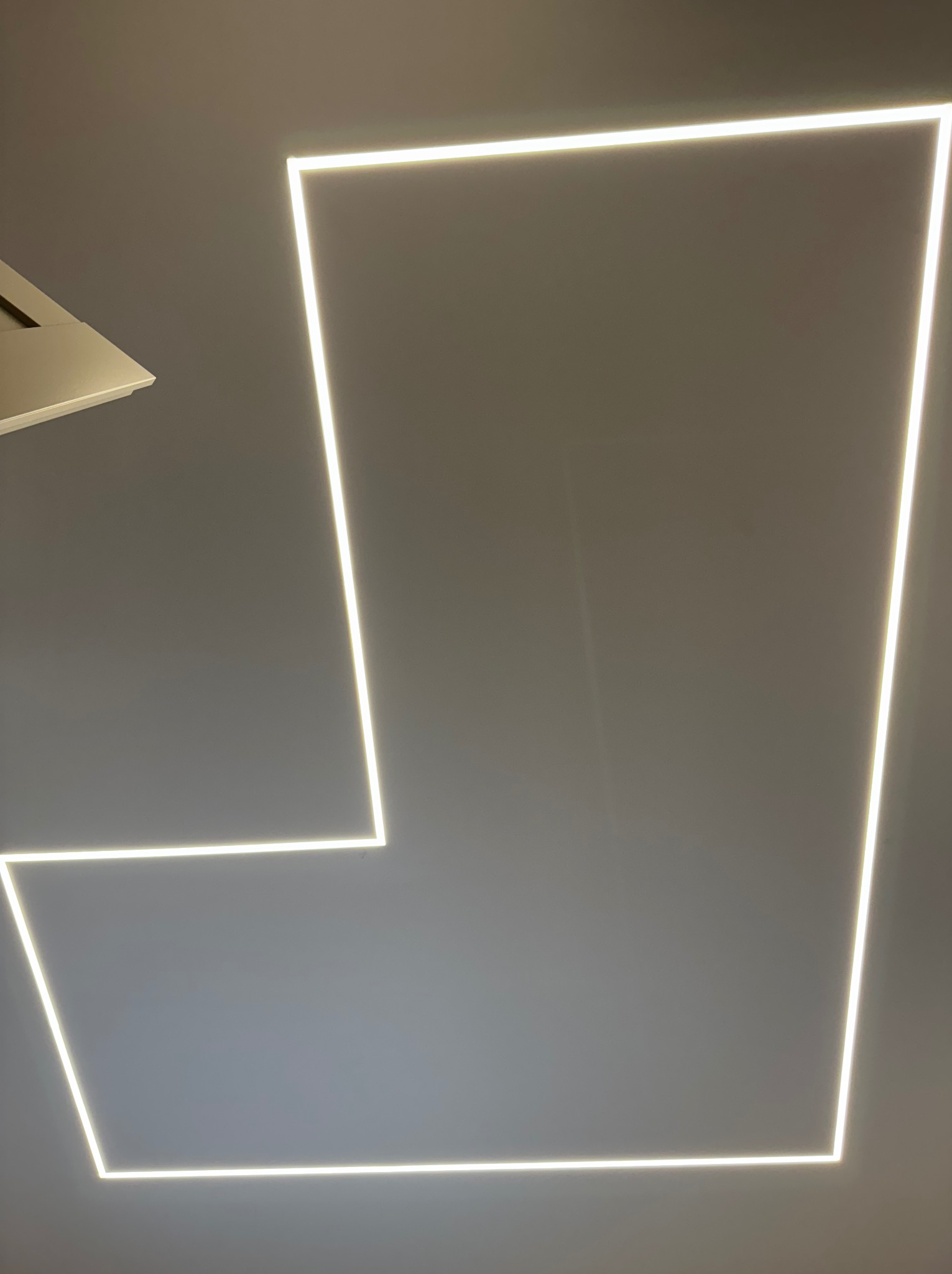 Nowoczesne oświetlenie sufitowe LED w kształcie nieregularnego czworoboku, z fragmentem wentylacji po lewej stronie. Minimalistyczny design, ciepłe światło.